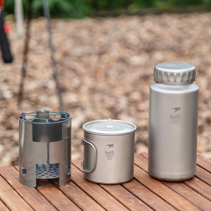 Keith Titanium Ti3070 wood stove, mug, and bottle displayed on a wooden table outdoors