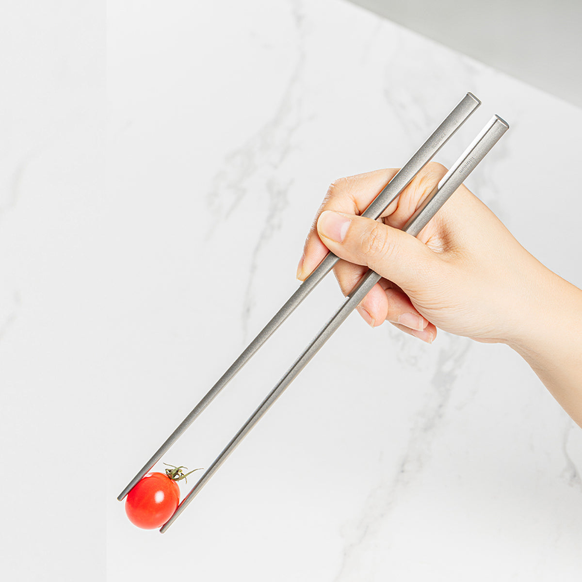 Hand holding Keith Titanium Ti5633 titanium chopsticks with a cherry tomato
